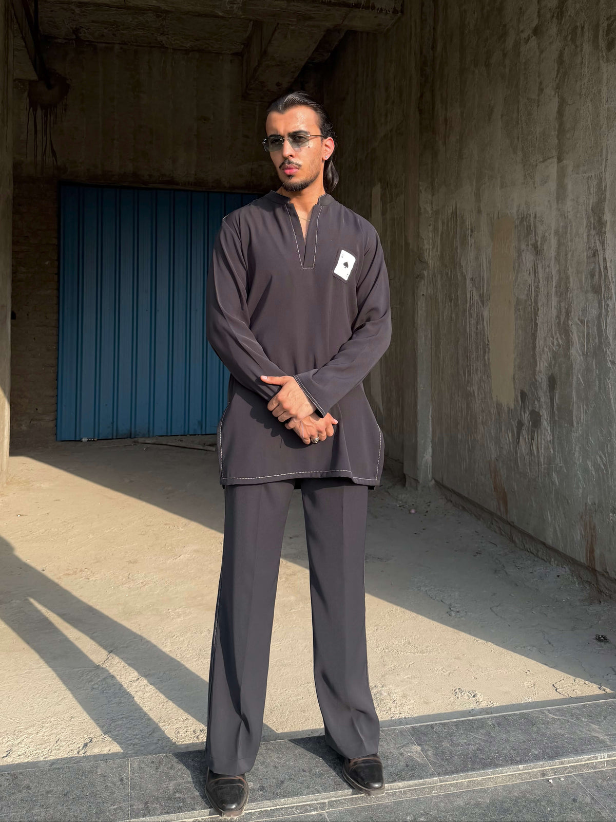 A man wearing a dark gray long-sleeve quarter-zip pullover and matching wide-leg trousers stands in a concrete structure with a blue corrugated metal door in the background.